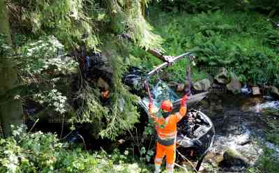 PKW-Hybrid stürzt mehrere Meter in Flussbett: Ersthelfer retten Fahrerin aus „Schwarzwasser“, aufwändige Bergung des Fahrzeugs, Hochvolt-Container auf Grund von Hybridfahrzeug angefordert: Feuerwehr im Einsatz, Ersthelfer stürzen sich Flussbett hinab und bringen Fahrerin ans Land